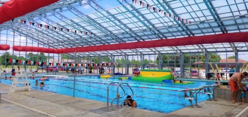 Indoor swimming pool with colorful floats, swimmers, and a lifeguard station under a glass roof.