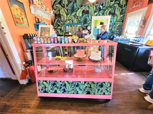 A colorful café display with a pink cart showcasing cakes and drinks, surrounded by vibrant tropical decor.