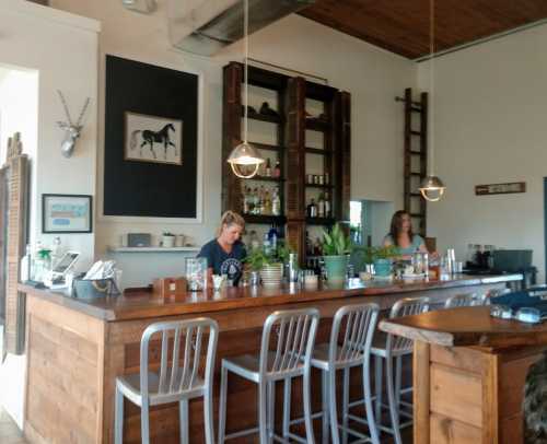 A cozy bar interior with wooden furniture, two bartenders, and shelves stocked with drinks and decor.