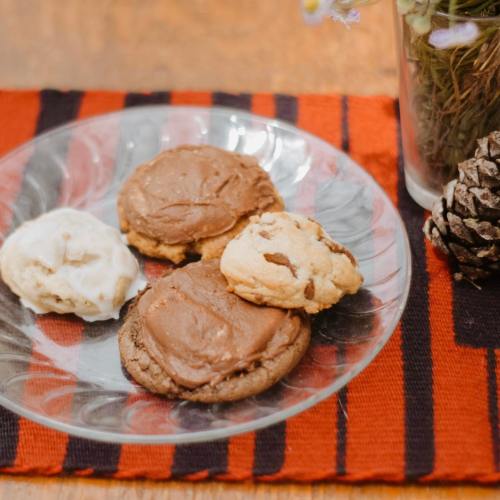 A glass plate with three cookies: two chocolate and one vanilla, on a striped orange and black cloth with a pinecone.