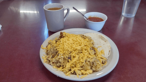 A plate of cheesy scrambled eggs with tortillas, a cup of coffee, and a bowl of salsa on a red table.