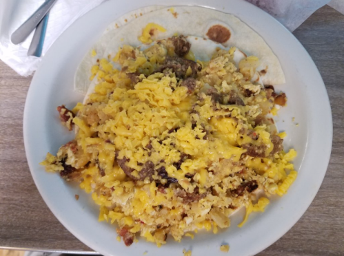 A plate of cheesy scrambled eggs with sausage and crispy bits, served over a tortilla.