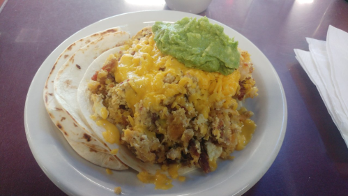 A plate of scrambled eggs with cheese, bacon, and guacamole, served with two soft tortillas.