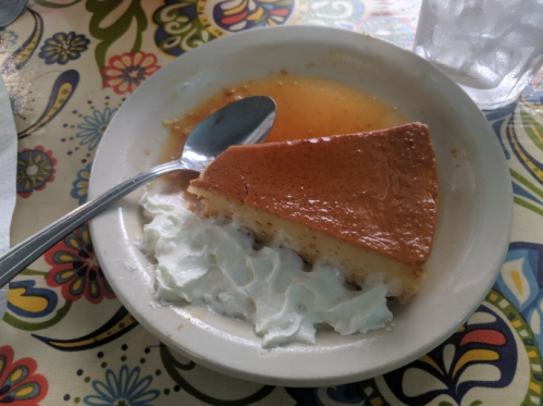 A slice of flan topped with caramel sauce, served with a dollop of whipped cream on a decorative tablecloth.
