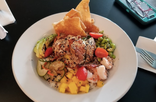 A colorful plate of grilled chicken, shrimp, rice, mango, edamame, and crispy tortilla chips, garnished with vegetables.