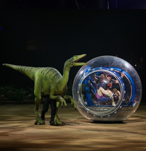 A performer in a dinosaur costume stands next to a futuristic vehicle with seated passengers in a dramatic stage setting.