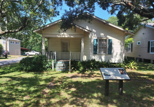 A small, light-colored house with blue shutters, surrounded by greenery and a sign in front on a sunny day.