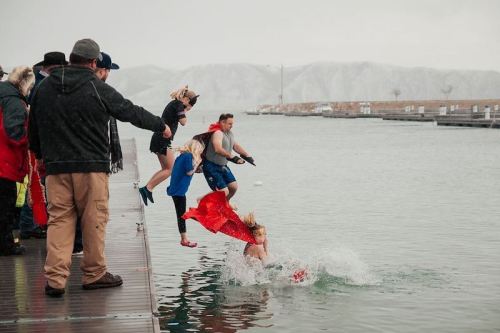 A group of people jumps into a chilly lake while onlookers watch from the dock, creating splashes in the water.