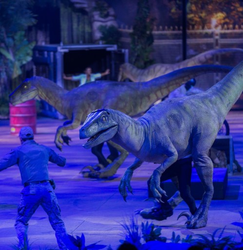 A performer interacts with lifelike dinosaur models in a vibrant, illuminated setting.
