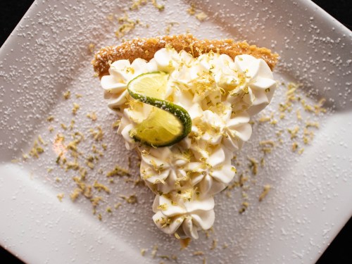 A slice of key lime pie topped with whipped cream, lime slices, and sprinkled with powdered sugar on a white plate.