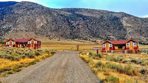 Two rustic cabins with red roofs sit on either side of a gravel road, surrounded by grassy fields and mountains in the background.