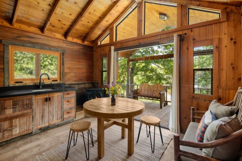 Cozy wooden cabin interior with a round table, chairs, and large windows overlooking a deck and greenery outside.