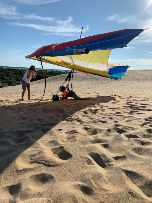 A person prepares a hang glider on a sandy beach, with another person lying underneath the glider.