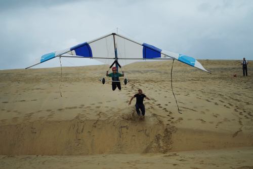 A person hang gliding above a sandy dune, while another person runs alongside on the ground.