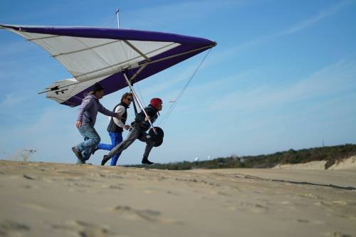 A person runs with a hang glider on a sandy beach, assisted by two others, under a clear blue sky.