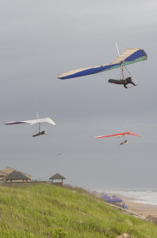 Three hang gliders soar above a grassy hill near the beach, with cloudy skies and distant tents visible.