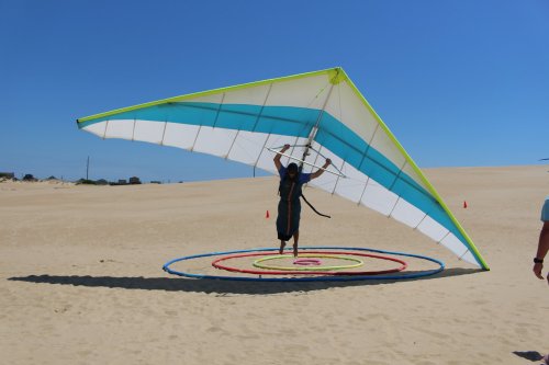 A person prepares to launch a hang glider on a sandy landscape under a clear blue sky.