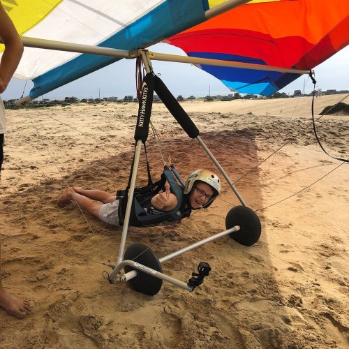 A child in a helmet gives a thumbs up while sitting in a hang glider on a sandy beach. Colorful wings are above.