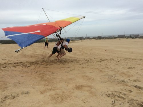 Two people are launching a hang glider on a sandy beach, with a cloudy sky in the background.