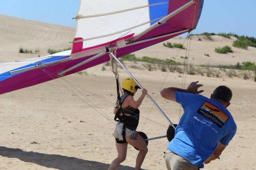 A person prepares for hang gliding on a sandy landscape, assisted by an instructor wearing a blue shirt.