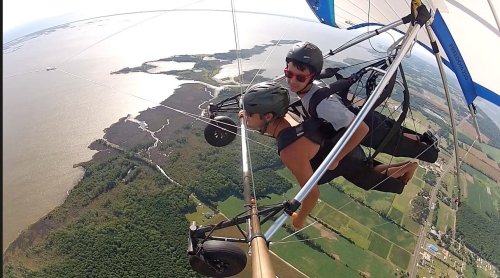 Aerial view of two people hang gliding over a scenic landscape with water and fields below.