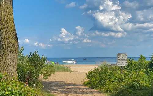 A sandy beach path leads to a calm lake with a boat in the distance under a blue sky with fluffy clouds.