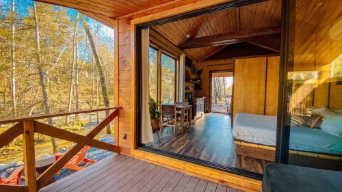 A cozy wooden cabin interior with large windows overlooking a forested area, featuring a bed and dining space.