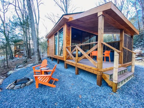 A cozy wooden cabin with a porch, surrounded by trees and gravel, featuring orange chairs and a fire pit.