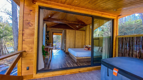 A cozy wooden cabin with large glass doors, revealing a minimalist bedroom and a hot tub on the deck.