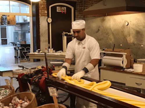 A candy maker in a white uniform shapes yellow taffy in a workshop with vintage equipment and wooden containers.