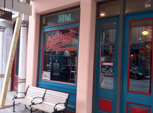A storefront with a colorful sign reading "La King's Confectionery," featuring an ATM and a bench outside.
