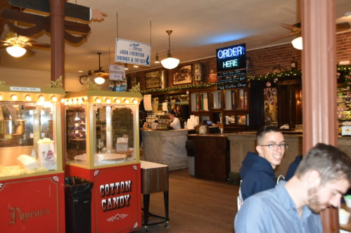 A vintage-style snack bar with popcorn and cotton candy machines, featuring a neon "Order Here" sign in a lively setting.