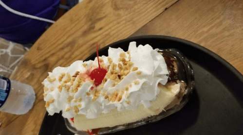A banana split topped with whipped cream, a cherry, and crushed nuts, served on a black plate.