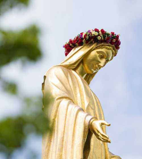 A golden statue of a woman with a floral crown, gazing downwards, surrounded by greenery.