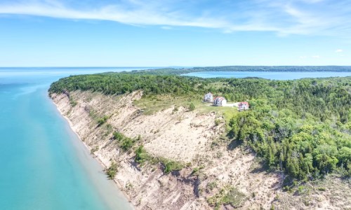 Aerial view of a coastal cliff with lush greenery and a house overlooking a calm blue lake under a clear sky.