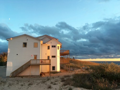 A modern two-story house on a sandy hill, with a wooden deck, against a backdrop of cloudy skies and a setting sun.
