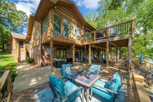 A spacious wooden deck with seating overlooks a serene lake, surrounded by trees and a charming house.