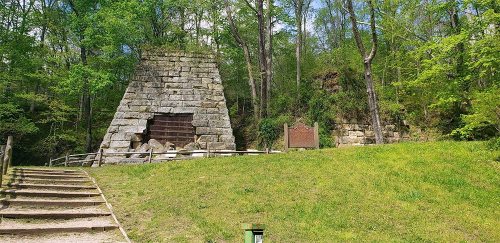 Stone structure resembling a pyramid, surrounded by trees and grass, with a sign nearby and steps leading up to it.