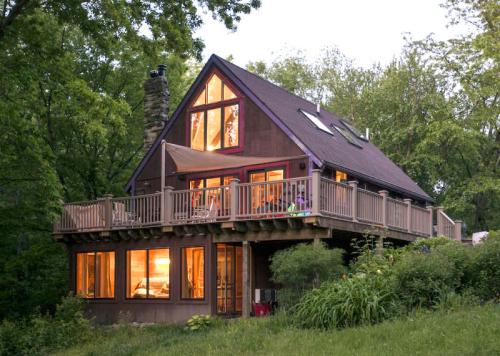 A cozy wooden house with large windows, surrounded by greenery, illuminated warmly at dusk.