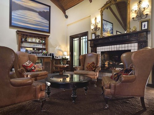Cozy living room with leather armchairs, a round table, a fireplace, and a bar area, featuring warm lighting and artwork.