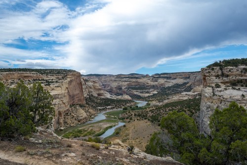 A scenic view of a canyon with a winding river, surrounded by rocky cliffs and green vegetation under a cloudy sky.
