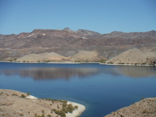 A serene lake surrounded by rocky hills under a clear blue sky. Reflections shimmer on the water's surface.