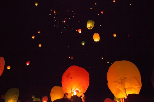 Colorful lanterns float into a dark night sky, illuminating the scene with warm hues of red, orange, and yellow.