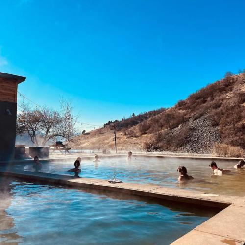 People relaxing in a steaming hot spring pool with a clear blue sky and hills in the background.