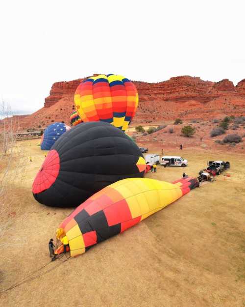 Colorful hot air balloons being prepared for flight in a grassy area with red rock formations in the background.