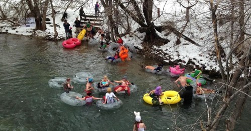 People enjoying a winter river scene, floating on colorful inner tubes amidst snowy trees and a chilly atmosphere.