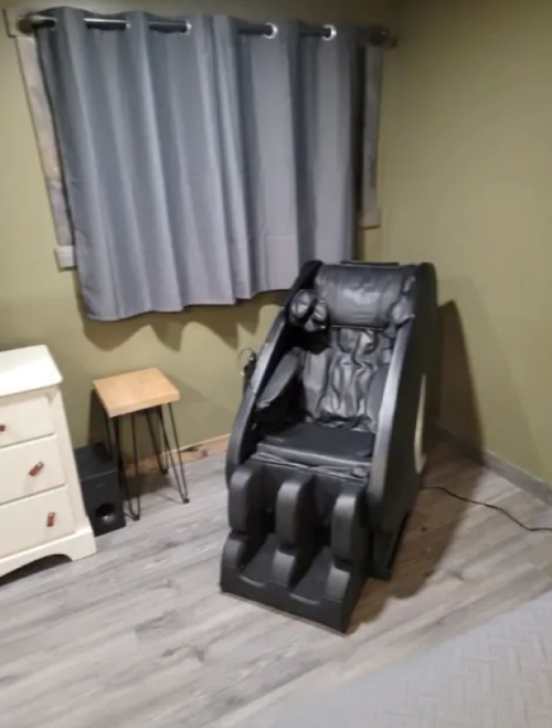 A black massage chair in a room with green walls, gray curtains, and a small wooden table nearby.
