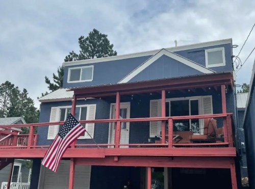 A two-story blue house with a red deck and an American flag hanging from the porch. Trees are visible in the background.