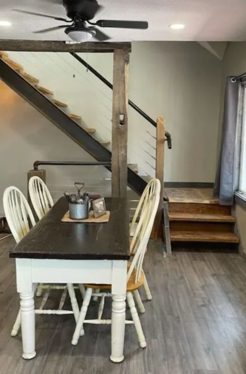 A cozy dining area with a wooden table and chairs, next to a staircase with a wooden railing and gray walls.