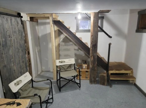 A basement with a staircase, wooden beams, and two folding chairs set up in a cozy corner.